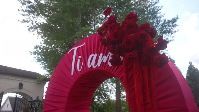 Low-angle magenta event arch topped with red roses and tassels against leafy outdoor venue, with arch, rose, flower, magenta detail, showing red, crimson, fabric, pleat context h