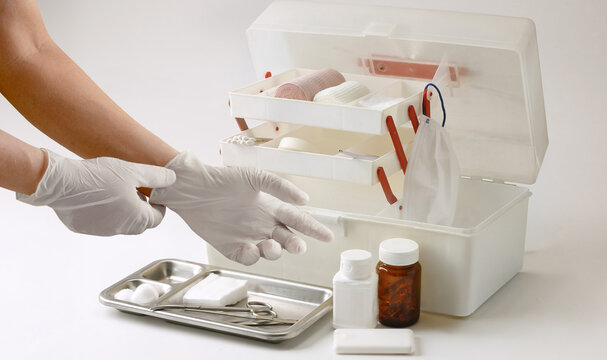 Healthcare professional wearing protective gloves preparing a first aid kit with medical supplies, bandages, gauze, surgical tools and face mask on a clean white background.
