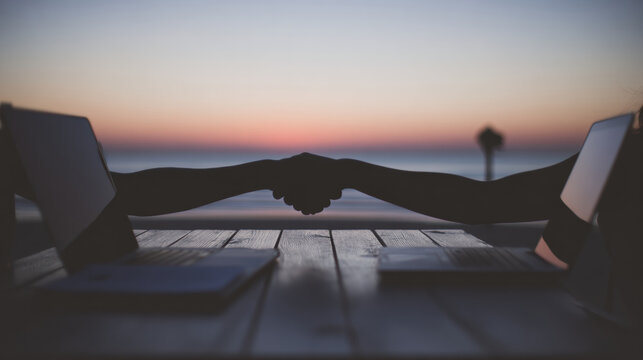 People shake hands during sunset at a beach with laptops on a table