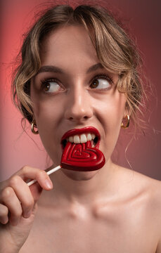 Playful woman biting heart-shaped lollipop with red lips looking sideways feeling flirty passion and desire