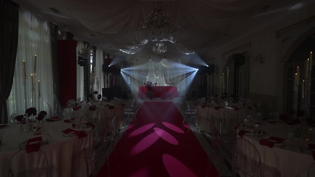 Converging spotlights illuminate red carpet aisle toward elevated head table in elegant banquet hall, with spotlight, beam, red, carpet detail, showing stage, headtable, banquet hall context