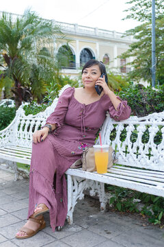 Woman in Purple Dress Talking on Phone on White Bench in Tropical Plaza