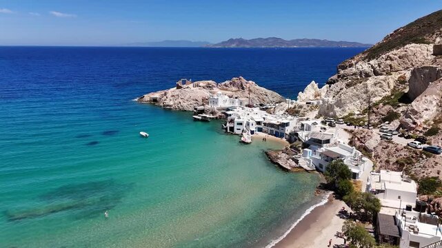 Firopotamos beach curves toward white houses as an aerial view advances along Milos, with turquoise shallows, a boat, rocky headland, parked cars and distant islands in bright sun.
