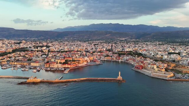 Chania Old Venetian Harbor and historic waterfront extend around the blue bay, with the Venetian breakwater, lighthouse, Crete mountains and dense cityscape, aerial shot.