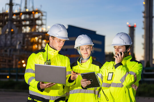 Oil refinery engineers team with laptop and tablet working at industrial plant at twilight, Petroleum plant worker and technician in safety gear using computer and radio communication in evening