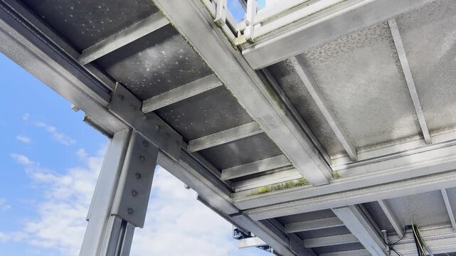 Low-angle view beneath a galvanized steel platform at a canoe sprint venue in Brandenburg, showing beams, bolts and modular structural construction against blue sky.
