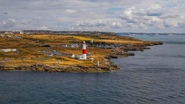 Aerial view of Portland Bill Lighthouse on the rocky coastline of the Isle of Portland, surrounded by grassy fields and the English Channel under a cloudy sky Portland, Dorset, United Kingdom.