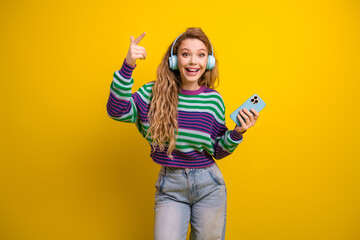 Young casual woman with headphones holding phone smiling against bright yellow background for...