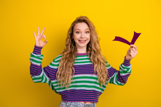 Young woman smiling in a colorful striped sweater holding a purple cutout against a yellow background showcasing fashion style and happiness