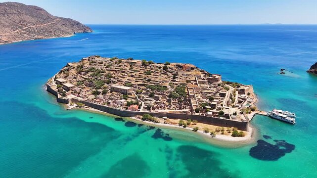 Kalydon fortress island recedes during an aerial pullback off Crete, revealing turquoise shallows, dark seabed patches, stone ruins, defensive walls, a small beach and docked ferry.