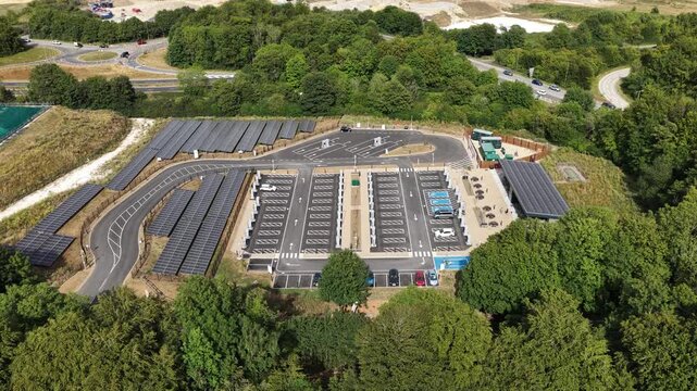 High aerial view orbiting a modern electric vehicle supercharging station, showcasing numerous charging stalls, solar panel arrays, and surrounding green trees providing clean, renewable energy