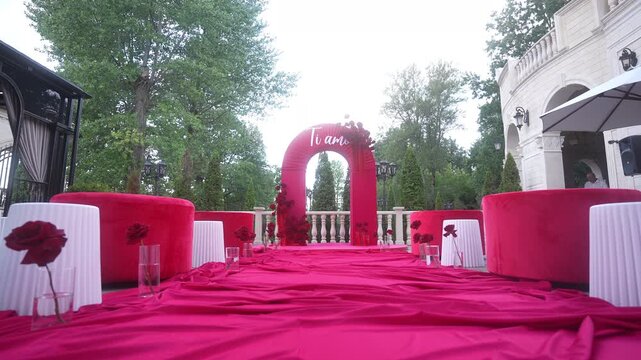Low-angle magenta ceremony arch on outdoor terrace with rose-lined aisle, velvet seating, stone balustrade, lanterns, pergola,and floral details creating elegant event setting