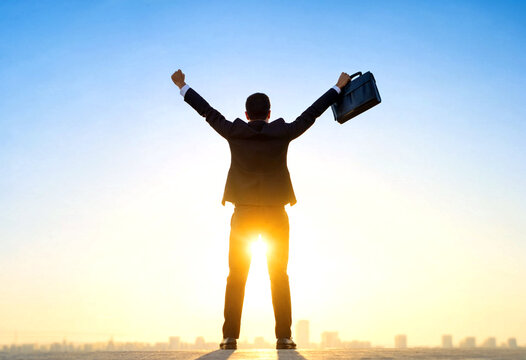 Successful businessman standing with arms raised holding a briefcase at sunrise over city skyline