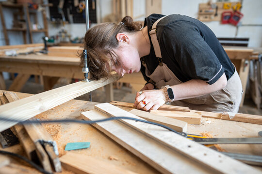 Passionate woman crafter at work in restoration workshop, working with wooden material at table, focused craftswoman preparing and examining parts for repair, construction and renovation in woodshop.