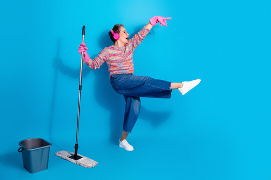 Joyful woman dancing while cleaning with a mop and bucket, listening to music in pink headphones and wearing rubber gloves on a bright blue background