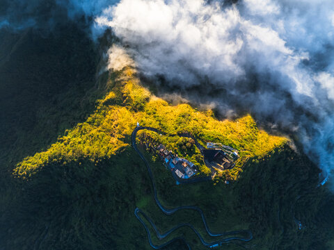 Aerial view of Mount Telomoyo with its winding access road, lush green mountain slopes, and thick clouds at sunrise in Kecamatan Ngablak, Jawa Tengah, Indonesia.