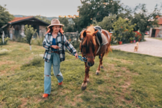 Blurred woman in a straw hat leads a saddled chestnut horse across a sunny farm yard, walking ahead with a relaxed smile. Rural lifestyle concept.