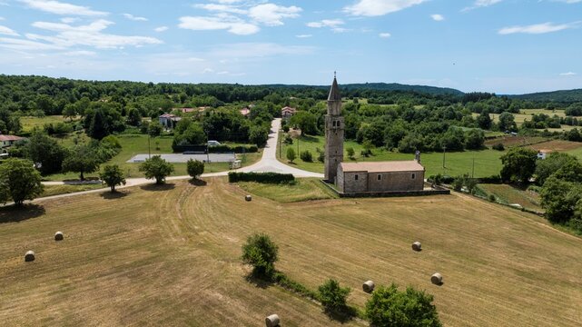 Aerial view of the historic stone church surrounded by rolling green fields, hay bales, and lush trees in Sterna, Istarska zupanija, Croatia.