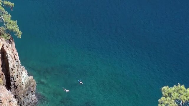 Two tiny figures navigate the mesmerizing blue-green ocean beneath an impressive, towering cliff. The tranquil natural beauty creates a perfect summer escape.
