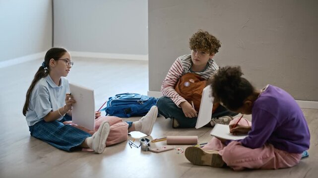 Multiethnic children sitting on floor of art gallery, drawing in sketchbooks and discussing school project. Perfect for elementary education promos and creative learning advertisements