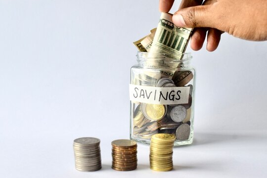 Hand placing US dollar banknote into glass savings jar beside three coin stacks.