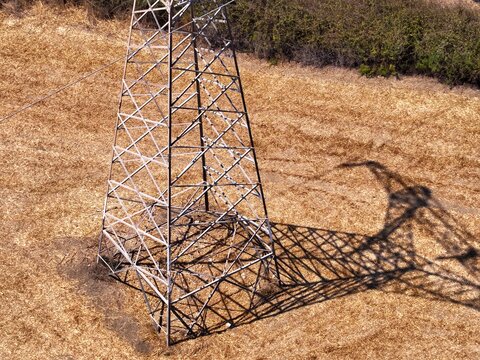 Aerial view of a metal electricity pylon casting a long shadow on a dry, golden field in Chania, Greece.