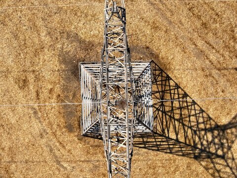 Aerial view of a metal electricity pylon casting a long shadow over dry golden fields in Chania, Greece.