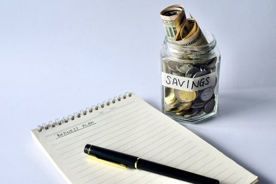 Angle view of a notebook inscribed with Business Plan next to a pen and a glass jar filled with coins and cash notes.