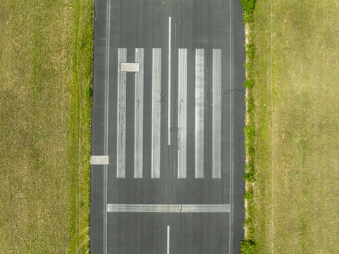 Aerial view of the historic Landebahn Neubiberg runway with faded white markings and cracks in the asphalt, surrounded by green grass fields in Unterhaching, Bavaria, Germany.