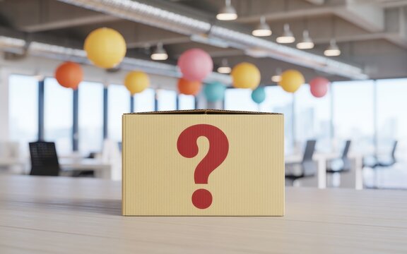 A cardboard box with a large red question mark printed on the front sitting on a wooden desk in a blurred modern office environment with colorful hanging paper lanterns