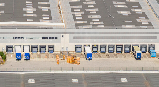 Aerial view of a large logistics warehouse with multiple loading docks, parked truck trailers, and stacked wooden pallets, showing freight distribution in Le Pouzin, Auvergne-Rhone-Alpes, France.