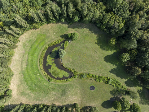 Aerial view of a crescent-shaped pond surrounded by lush green grass and dense forest in Aschheim, Bavaria, Germany.