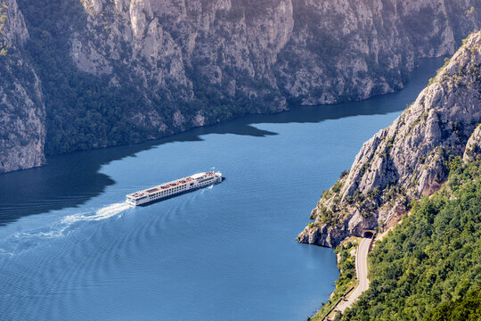 A white river cruise ship glides through a dramatic limestone Iron Gates canyon gorge, dwarfed by towering cliffs above a deep blue Danube river in the Balkans. Travel in Djerdap concept.