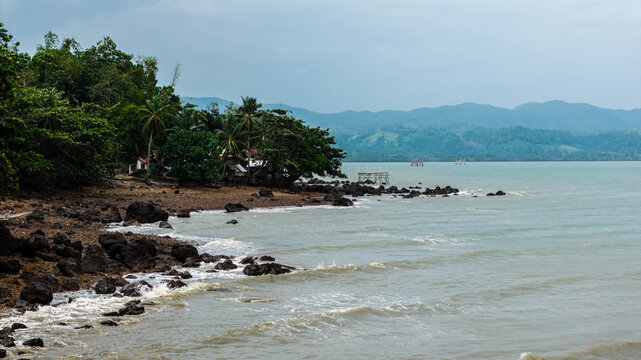 Aerial view of the rocky coastline with lush palm trees and vegetation along the shore, calm sea waters, and distant mountains in Barotac Viejo, Western Visayas, Philippines.