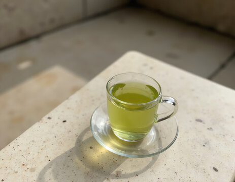 Clear glass cup of hot green herbal tea on saucer with natural sunlight and harsh shadows on stone surface