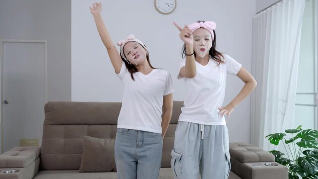 Two young Asian women having fun with a beauty routine, wearing facial sheet masks and pink headbands while sitting on a sofa. Happy friends enjoying a home spa and skincare self-care together.