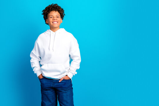 Smiling teenage boy in white hoodie and jeans standing confidently with hands in pockets against a bright blue background, casual youth portrait and lifestyle concept