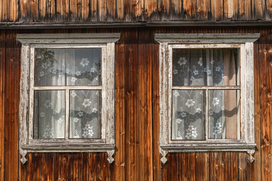 Facade of this old wooden house is decorated with two windows in peeling white carved frames, lace tulle curtains behind glass and boards darkened by time. Authentic rustic aesthetic