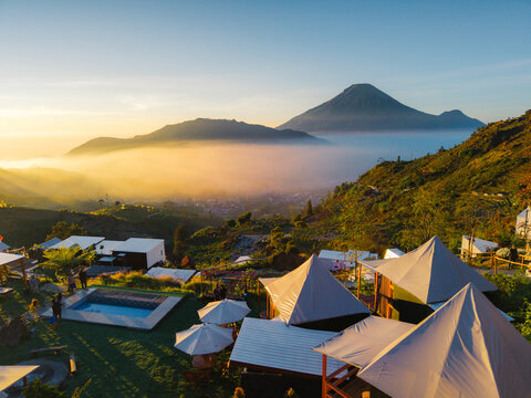 Aerial view of Sikapuk Hill and Villa Dieng featuring luxury glamping tents and a swimming pool set against a misty mountain landscape during golden hour in Indonesia.
