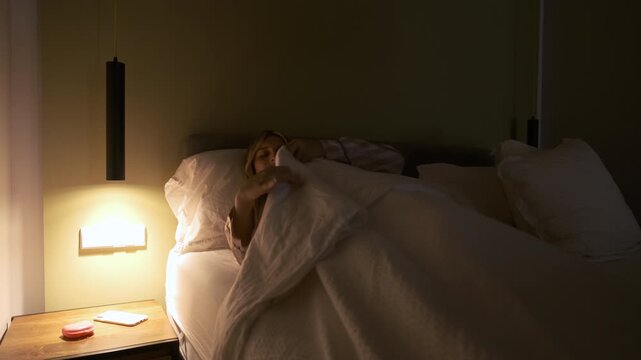 Young transgender woman in nightwear putting on duvet while lying in bed as she prepares herself for sleep in bedroom.