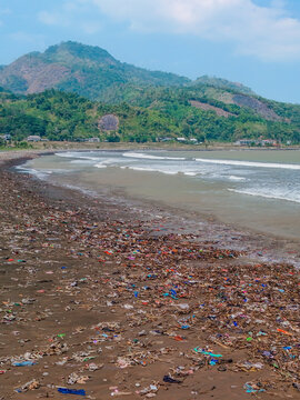 Aerial view of a beach heavily polluted with plastic waste and debris, with rolling waves and lush green mountains in the background, Indonesia.