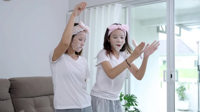 Two young Asian women having fun with a beauty routine, wearing facial sheet masks and pink headbands while sitting on a sofa. Happy friends enjoying a home spa and skincare self-care together.