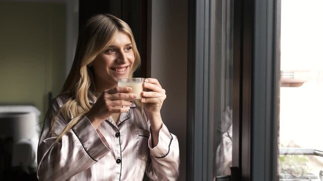 Thoughtful transgender woman in nightwear drinking fresh coffee as she looks through window while standing at home