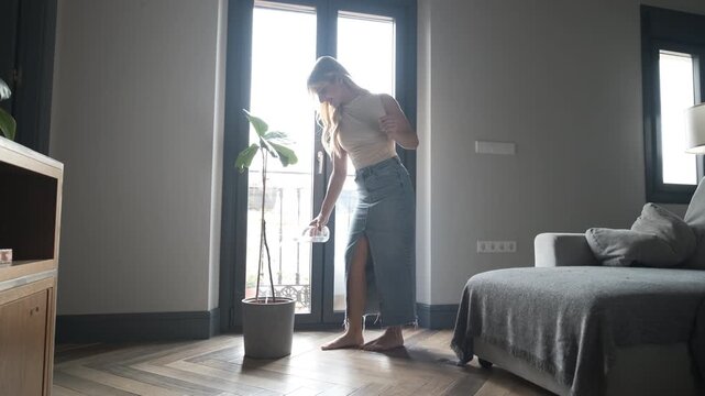 Full body of young transgender woman in casual wear pouring water with glass bottle in potted plant while standing on hardwood floor at home