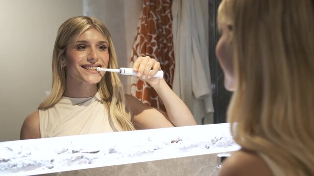 Reflection of blond female transgender brushing teeth with electric toothbrush in bathroom mirror at home
