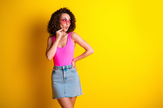 Young woman in pink top and denim skirt looking surprised with sunglasses against a vibrant yellow background, playful fashion portrait conveying fun, retro style and summer vibes