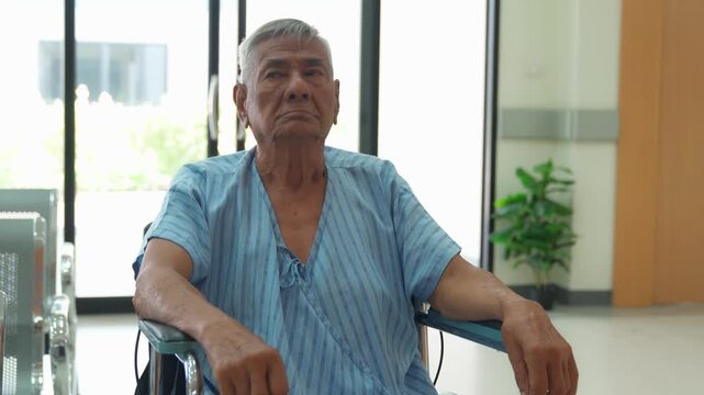 An elderly male patient sitting in a wheelchair wearing a hospital gown and orthotic footwear.