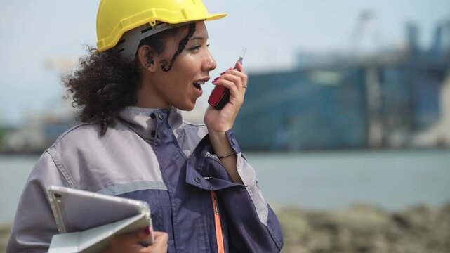 Female marine engineer reviewing clipboard before using walkie talkie at seaport, cargo vessel operation, shipping logistics, port inspection, maritime communication.