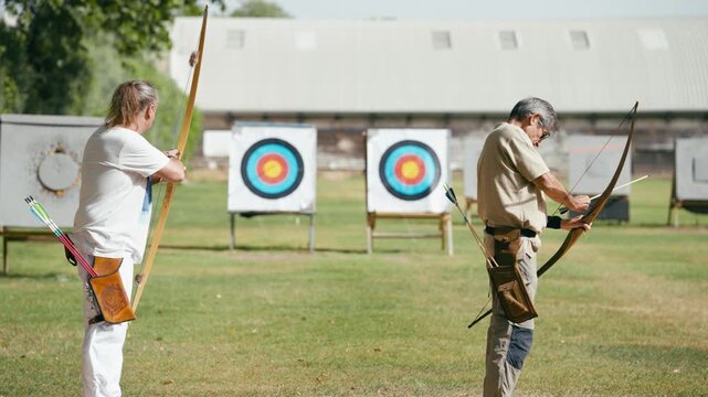Two older men practicing archery outdoors. An archer shooting an arrow from a traditional bow, hitting the target in the background