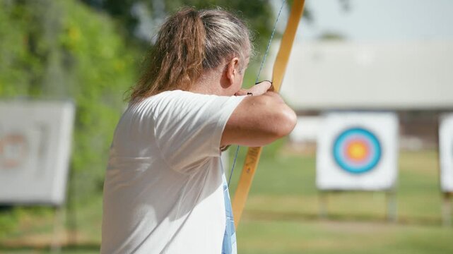 Focused mature man aiming and shooting an arrow with a traditional wooden longbow during an archery competition in an outdoor shooting range
